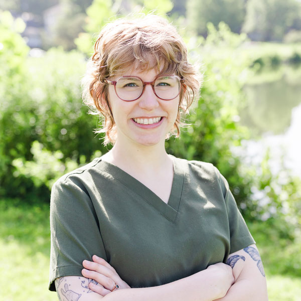 Zoe, a Certified Ophthalmic Assistant, stands by a pond wearing glasses and a green shirt, enjoying the tranquil scenery