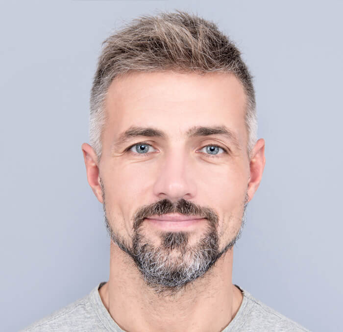 A bearded man wearing a grey shirt, looking confidently at the camera with a neutral background