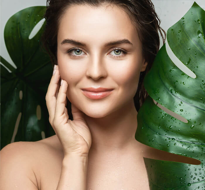 A woman with striking blue eyes stands against a backdrop of vibrant green leaves